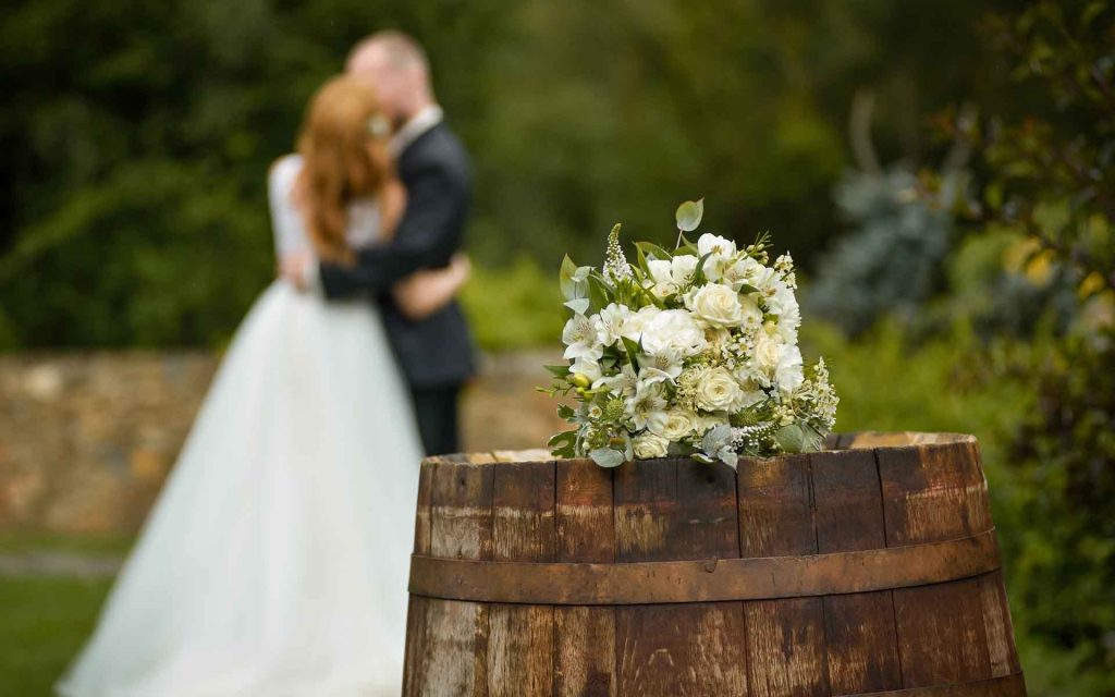 Bride and groom kiss in background with a flower bouquet on a barrell.